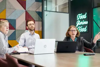 Three people sit in a meeting room with laptop looking at a screen